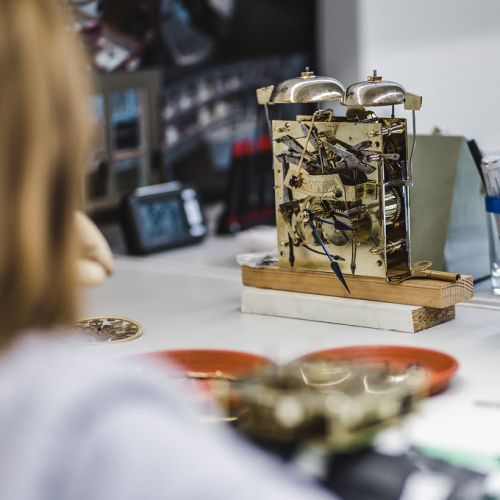 Am Arbeitstisch in der Werkstatt steht das freigelegte Uhrwerk einer Großuhr.
